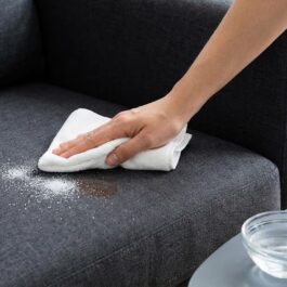 Main utilisant un chiffon microfibre blanc et une solution nettoyante pour tamponner une tache de sel sur un divan en tissu.